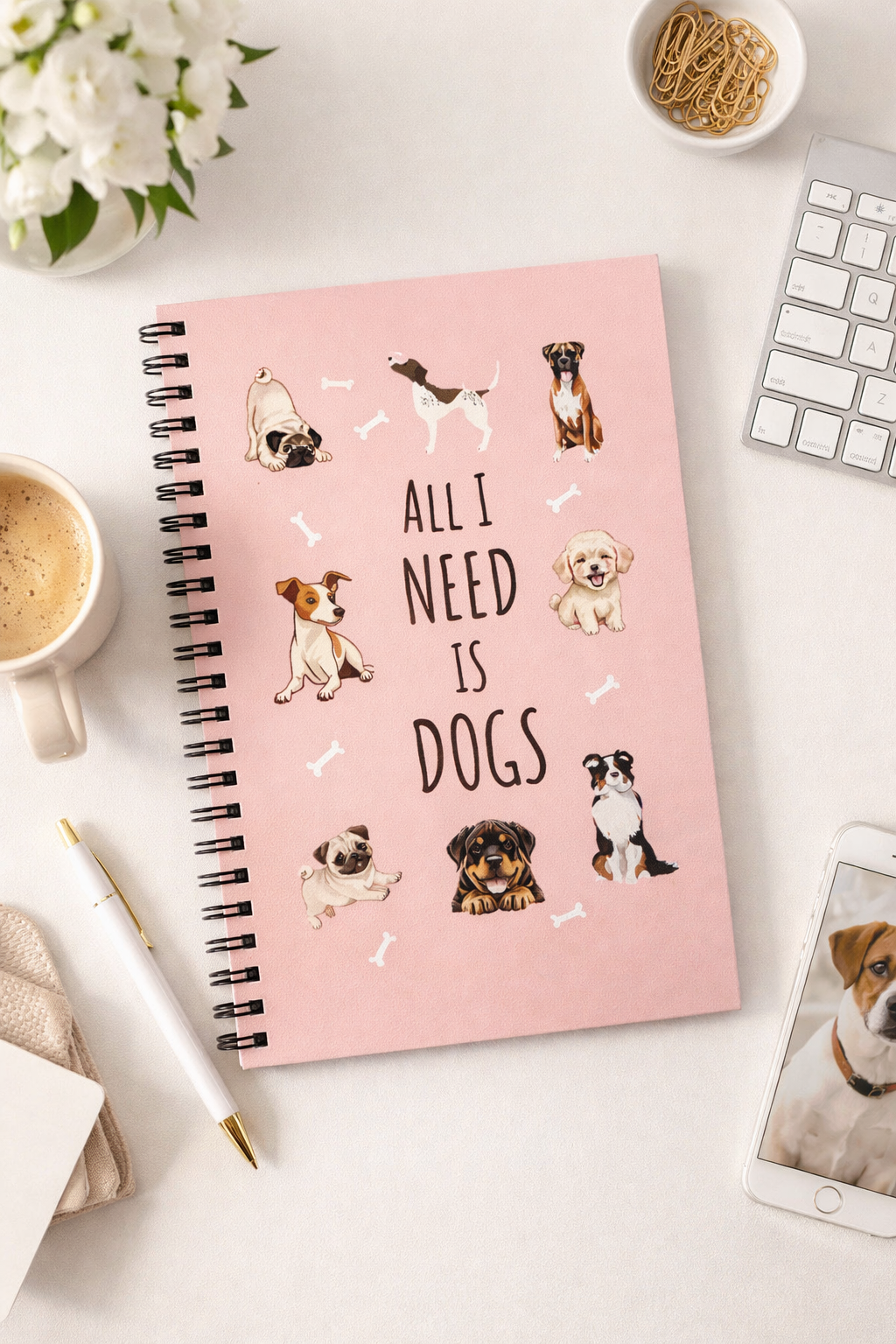 Pink notebook with dog illustrations and text 'All I Need is Dogs' on a desk with coffee and flowers.